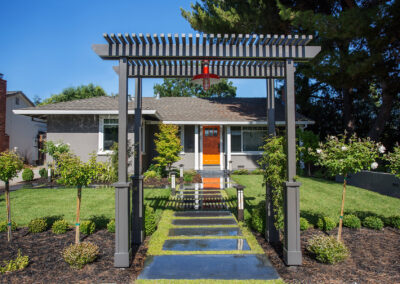 craftsman style pergola over a landscaped path leading to a gray house with orange front door